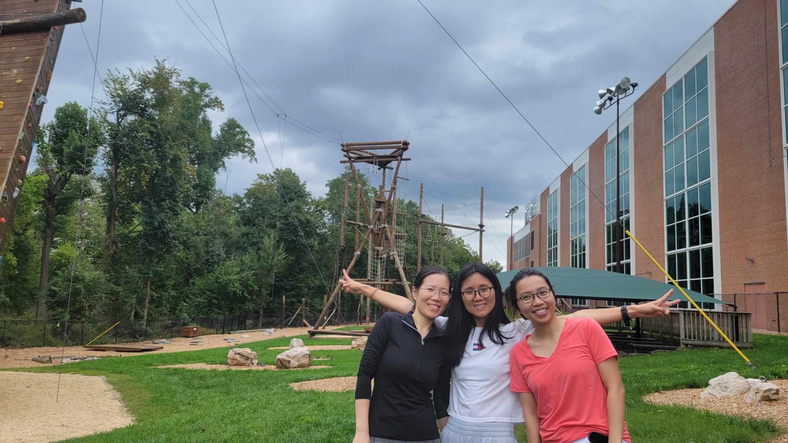 Three students smiling at the camera in front of an outdoor climbing wall